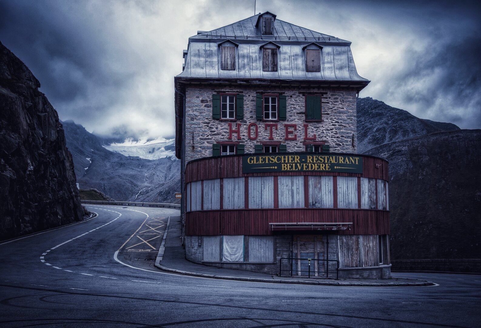 Ein altes Hotelgebäude, auf dem das Wort „HOTEL” in roter Schrift hervorsticht, liegt an einer kurvenreichen Straße in einer bergigen Landschaft. Das Hotel hat eine rustikale Steinfassade und grüne Fensterläden sowie ein Schild für ein Restaurant. Wolken bedecken den Himmel und verleihen der Szenerie eine zusätzliche Atmosphäre.
