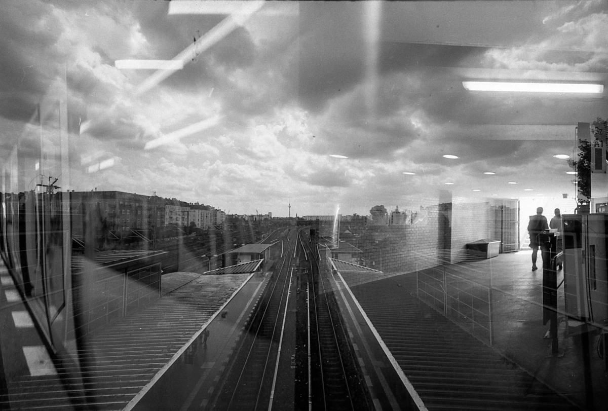 doppelt belichtetes schwarzweißfoto, wir sehen ein gleisbett, von einer brücke fotografiert, im hintergrund der fernsehturm und rechts und links bahnsteig, ein teil einer sbahn, menschen.
