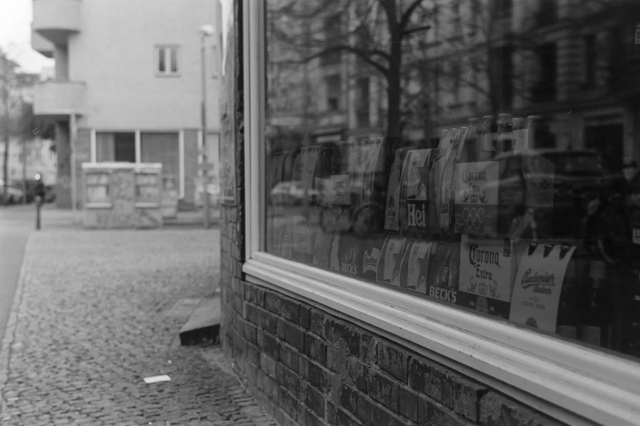 ein schwarzweißes bild, wir sehen eine große schaufensterscheibe, dahinter diverse sechserträger bier. der blick schweift zum hintergrund, da sehen wir gerade so noch ein mehrfamilienhaus.