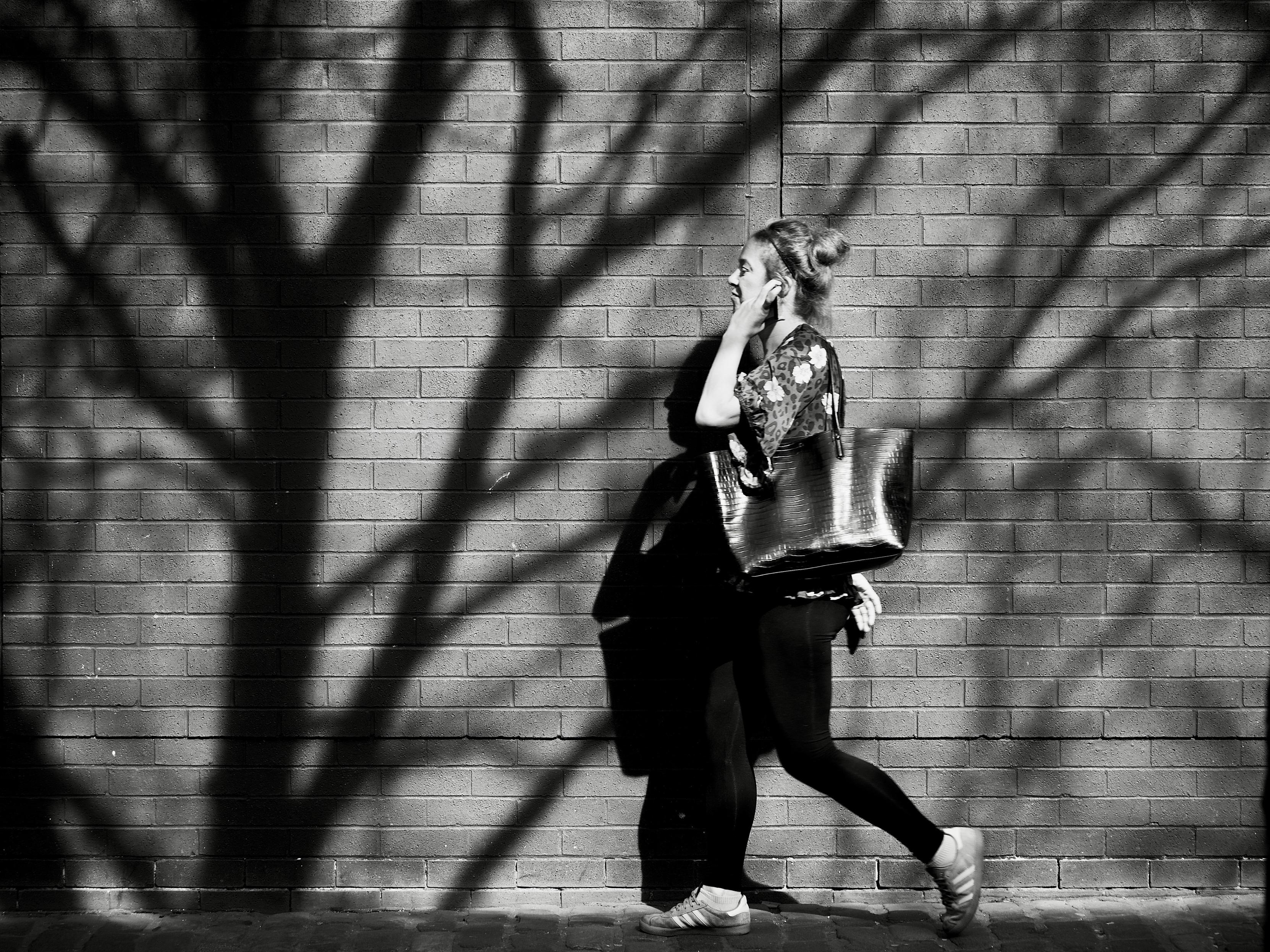 A young woman with her bag slung over her shoulder and her left hand to her ear is caught in a patch of bright sunlight on a city street lined with trees and on the wall immediately next to her are the dark, shadowed branches of a tree reaching out like tentacles.