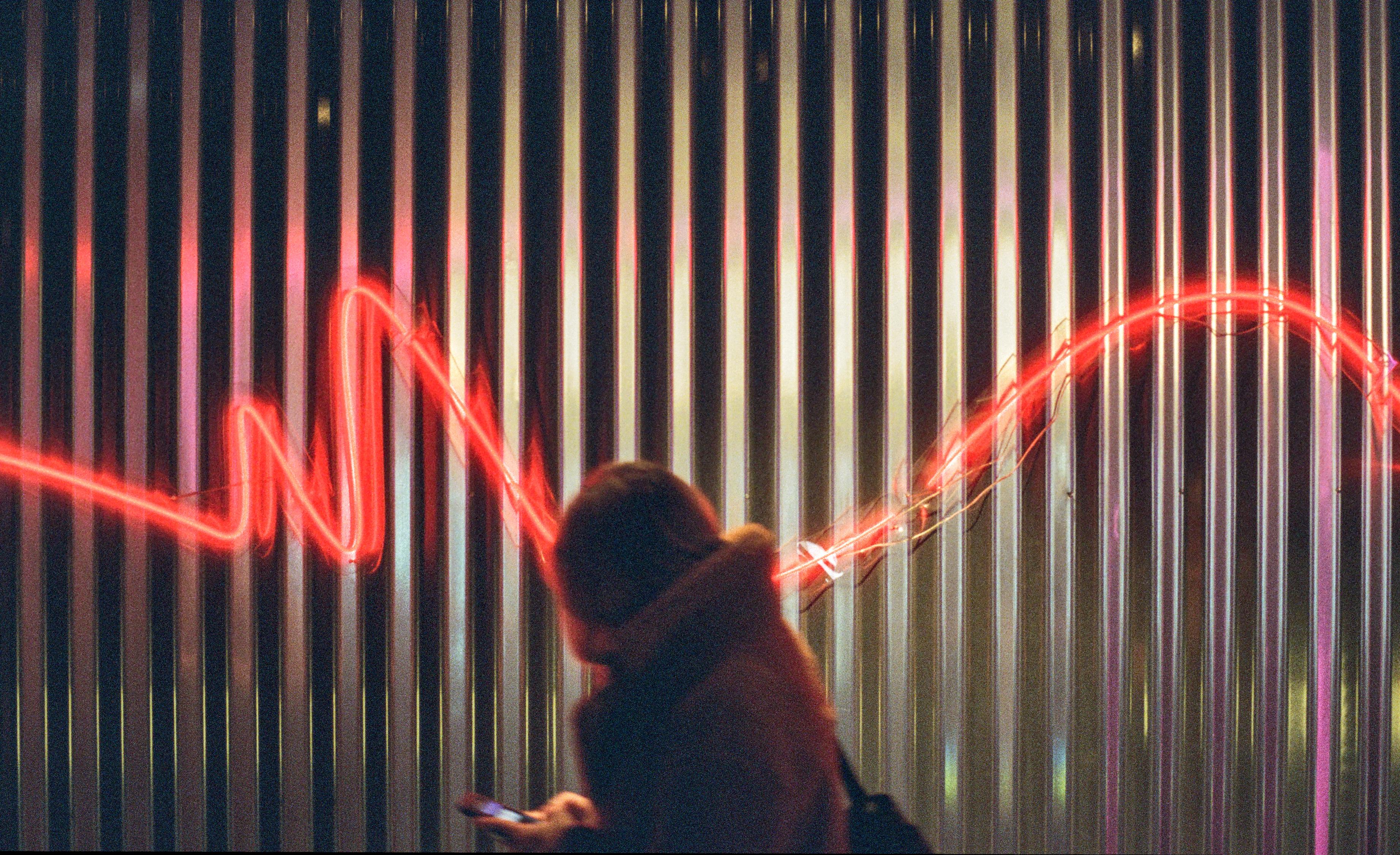 A woman walking past a wall of vertical metallic stripes, slightly blurred in motion. She looks down at her phone as she moves, her figure softened by the movement. Behind her, a vivid red neon line snakes across the scene like a glowing waveform, casting a warm, electric glow that contrasts with the cool-toned background. The image feels cinematic and dynamic, with motion blur emphasizing the fleeting, in-between moment.