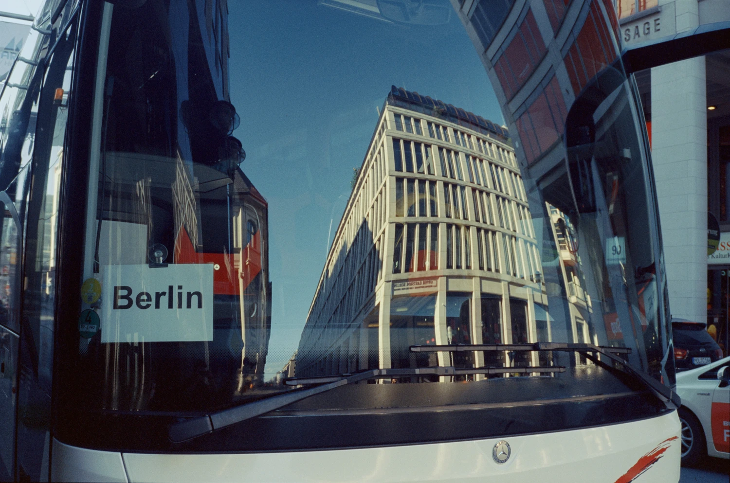 wir blicken in eine spiegelnde frontscheibe eines reisebus und sehen moderne architektur, etwas gewölbt. im bus ein schild auf dem Berlin steht.