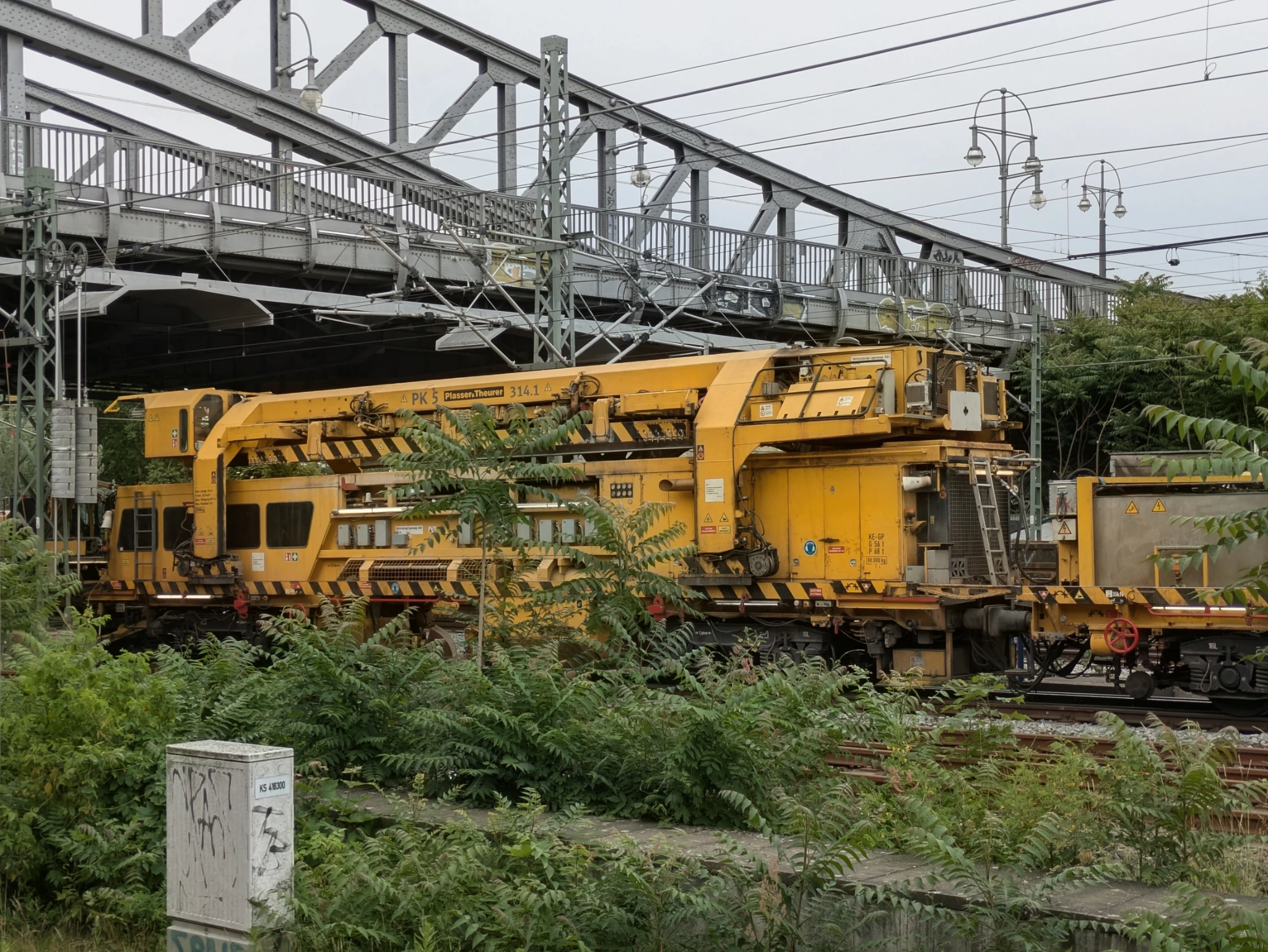 eine monströse gelbe maschine steht auf den gleissn einer Stahlskelettbrücke, viel gewächs drumherum